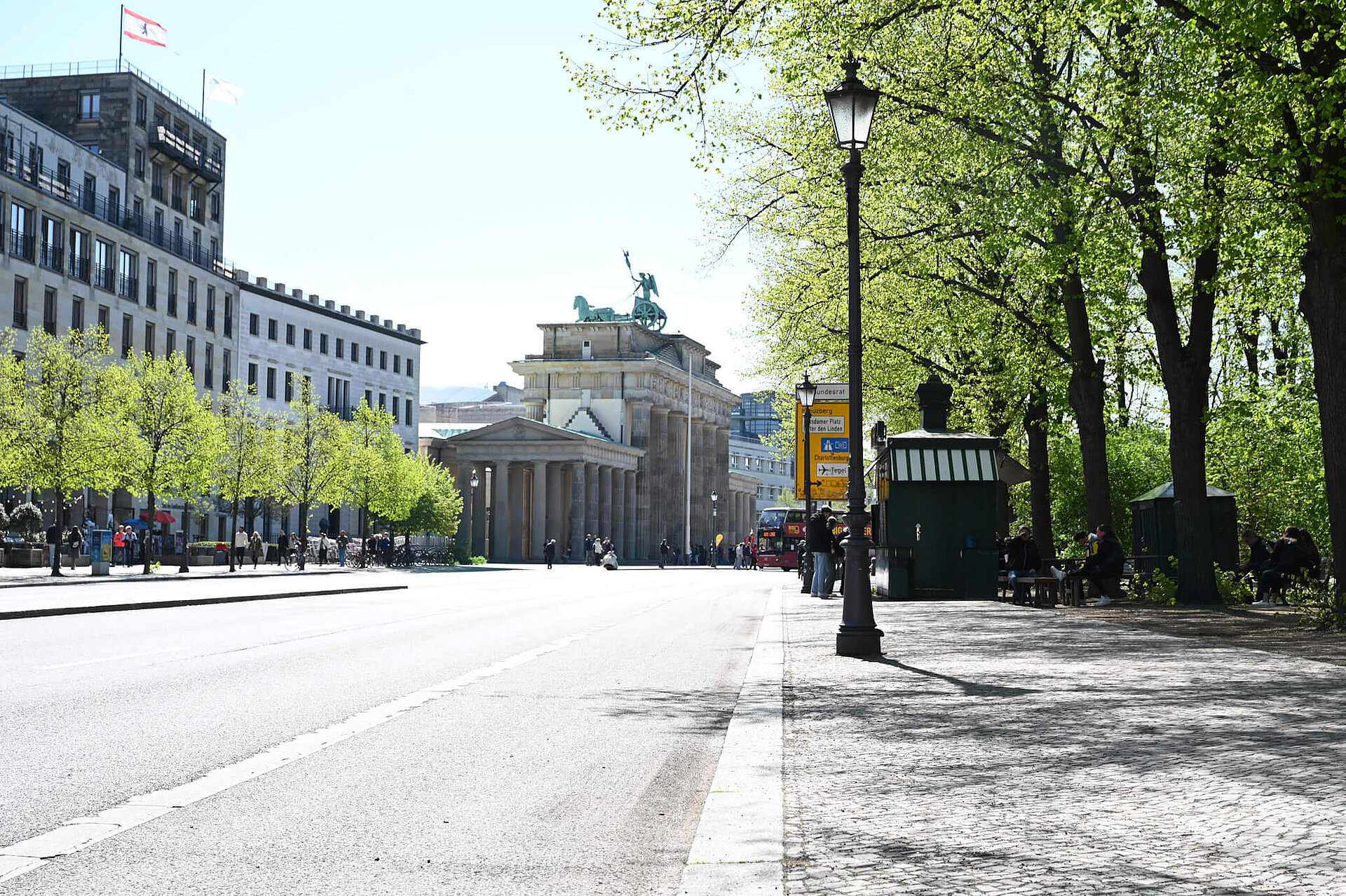 A view down the street towards the Brandenburg Gate. Green trees and a newspaper kiosk by the roadside.