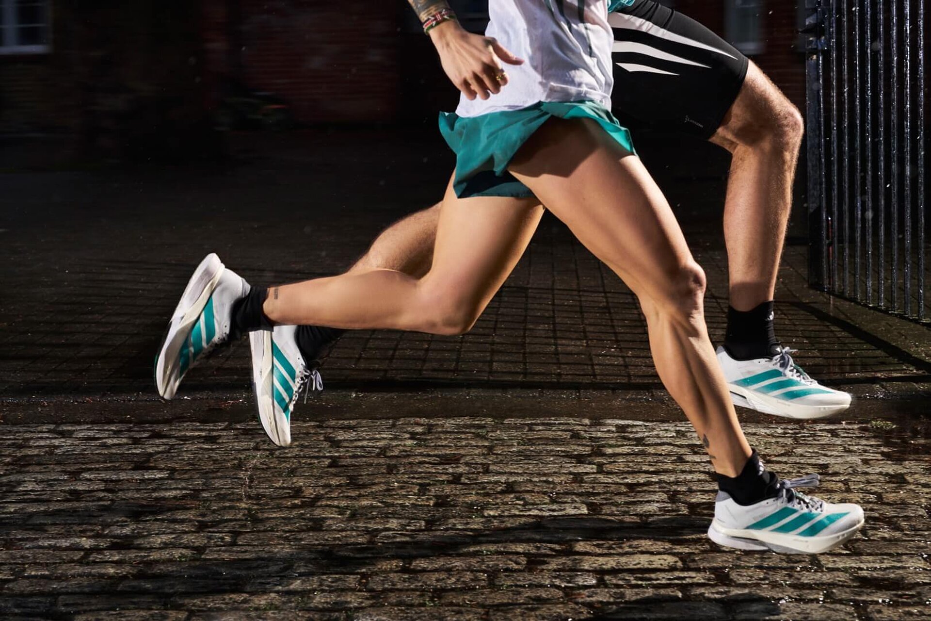 Two runners wearing white adidas shoes with green stripes are running across cobblestones. Only their legs are visible.