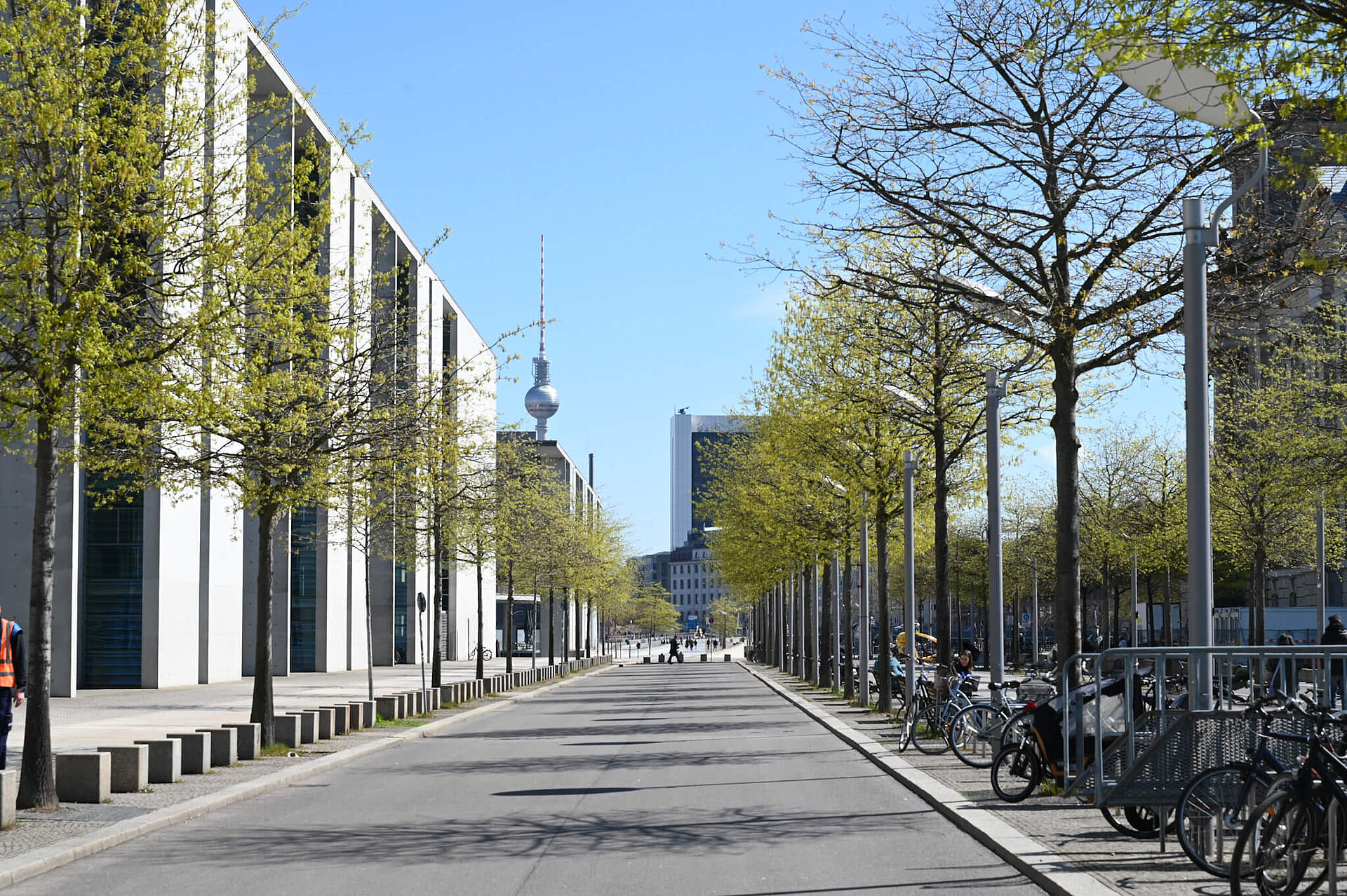 An avenue lined with young trees. On the left is the Paul-Löbe-Haus; on the right, a small section of the Reichstag building is visible. The TV Tower can be seen in the background.