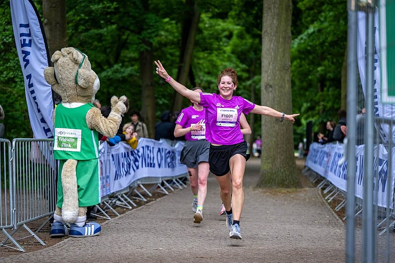 A woman wearing a T-shirt from the 40th VITAMIN WELL Women's Race Berlin runs toward mascot Frido with a smile to high-five him.