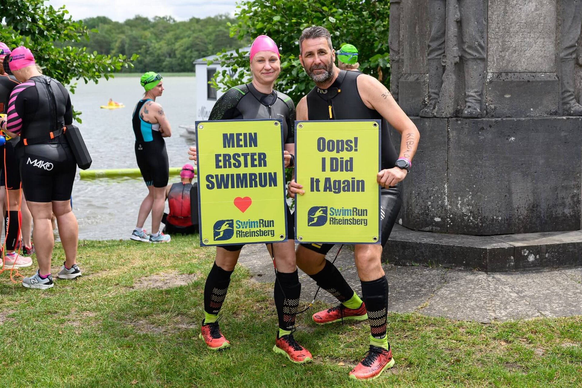Laura hält ein Schild mit Aufschrift "Mein erster SwimRun" und Herz-Emoji in die Kamera. Auf Philips Schild steht "Oops! I did it again".