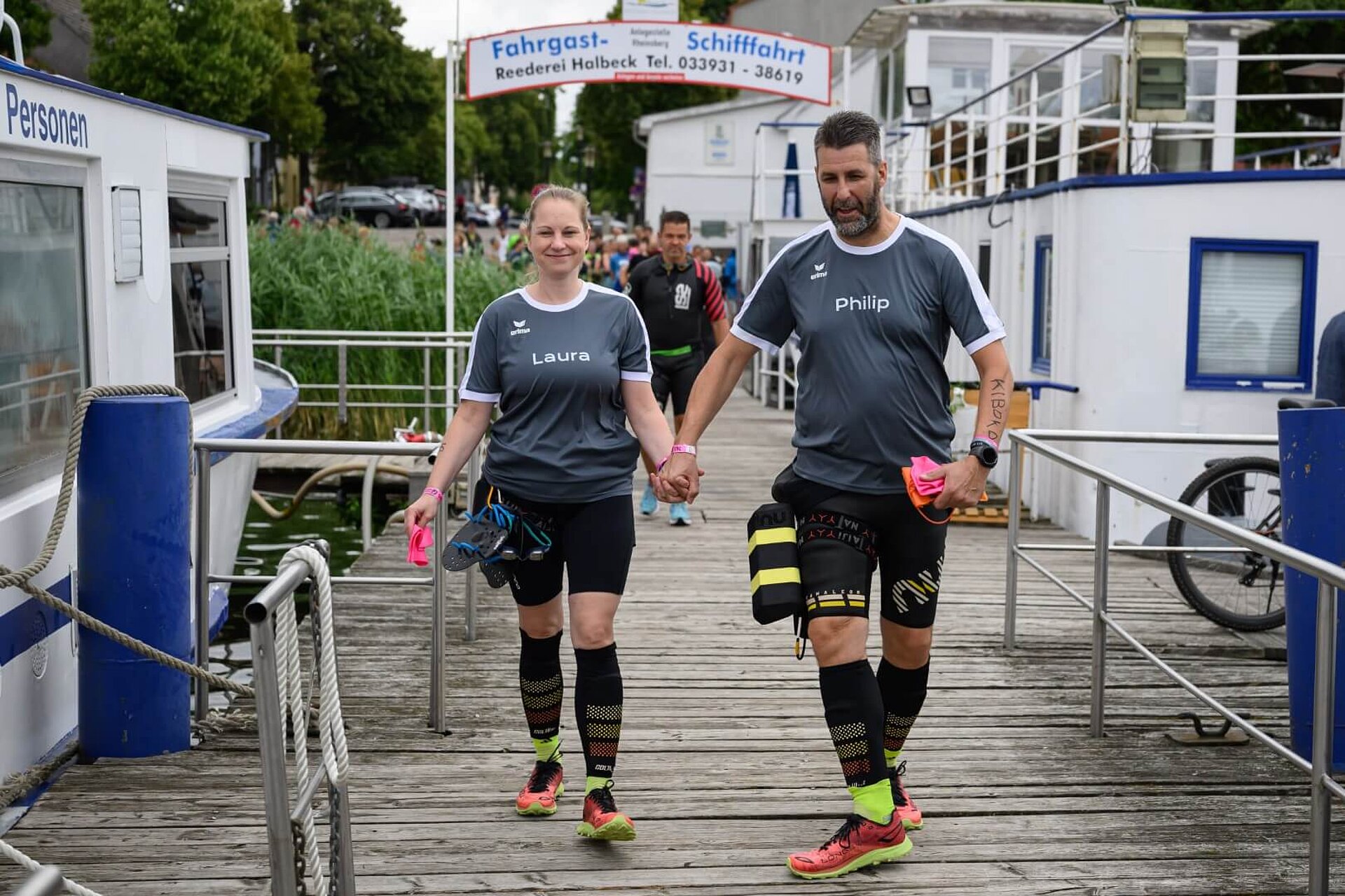 Laura und Philip in SwimRun-Ausrüstung gehen Hand in Hand auf einem Holzsteg.