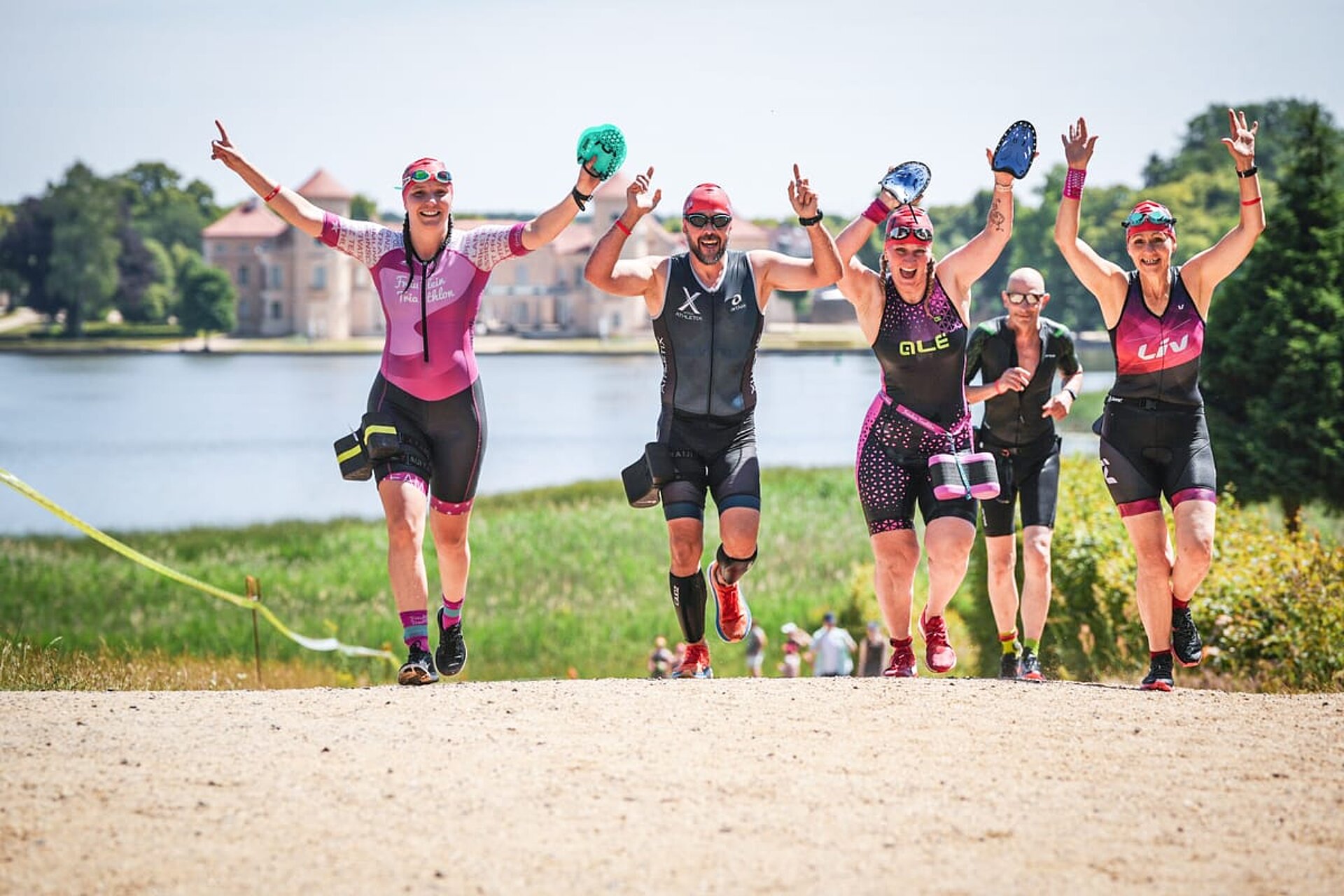Vier laufende Teilnehmende in Neopren mit Swimrun-Paddles und Pull Buoys strecken jubelnd ihre Arme in die Luft.