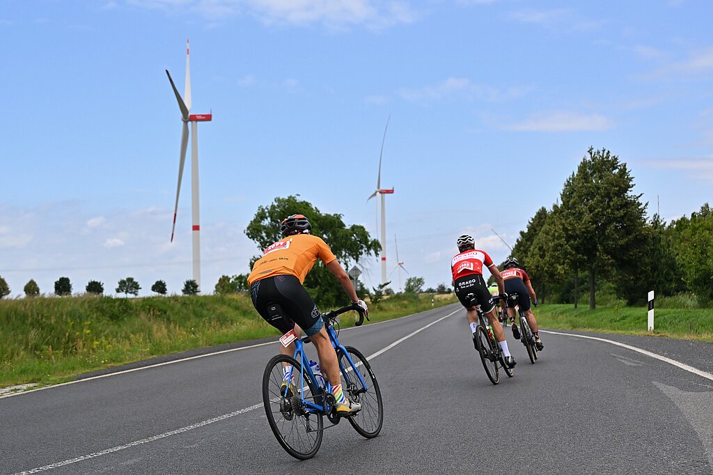 Radfahrer bremsen in einer Kurve auf der Landstrasse, Windkrafträder im Hintergrund © SCC EVENTS / Petko Beier