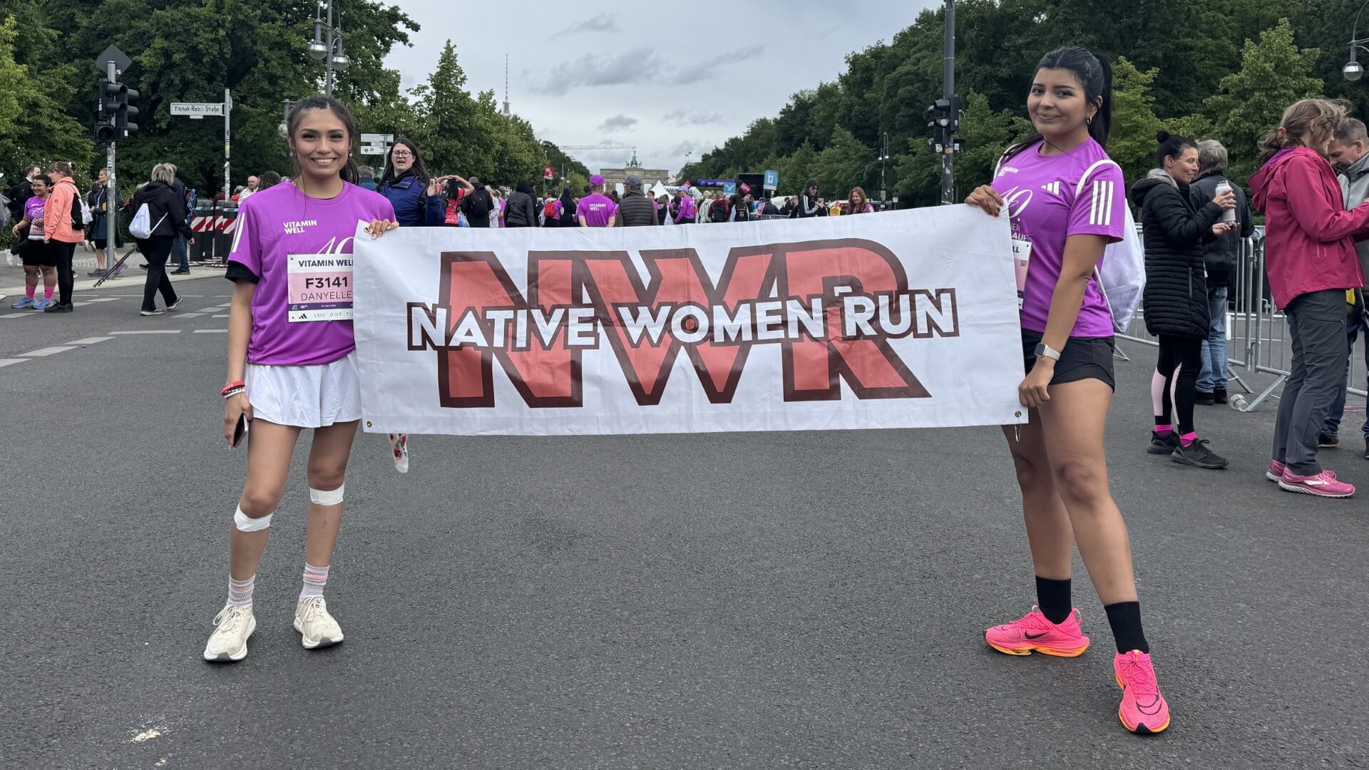 Zwei Läuferinnen halten lächelnd ein Banner mit der Aufschrift "Native Women Run" und der Abkürzung "NWR".
